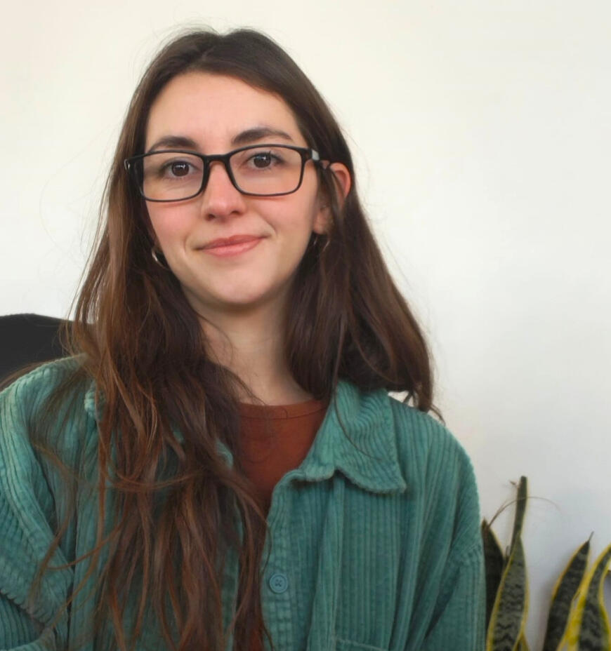 Picture of Gabriela Palachi, wearing a green shirt and black framed glasses. The background is white with some plants.